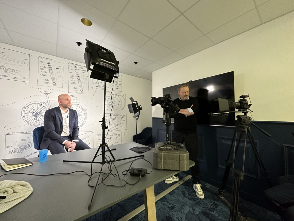 La personne interviewée lit son texte sur un prompteur face à lui. Une personne sur le côté supervise le tournage.