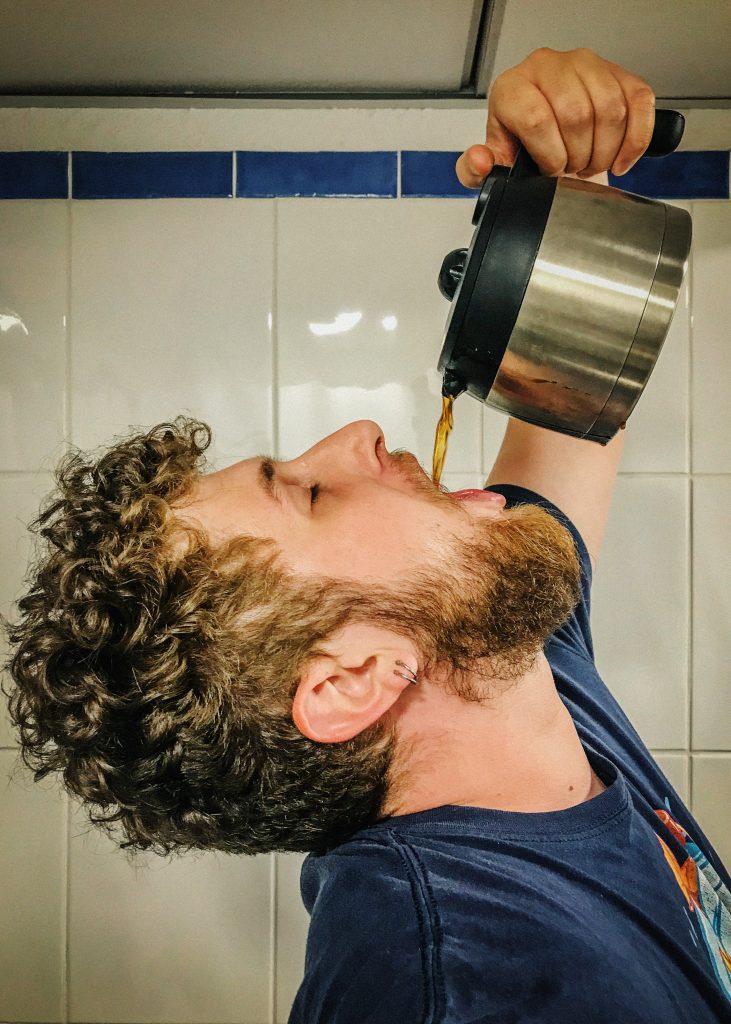 Un homme de côté qui tient une cafetière en l'air dont le contenu se déverse dans sa bouche.